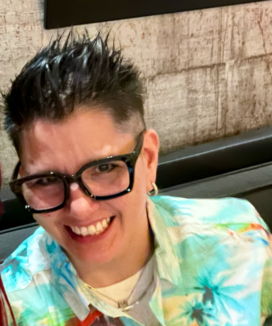 Canne, smiling with short spiky hair and glasses, wearing a colorful patterned shirt
